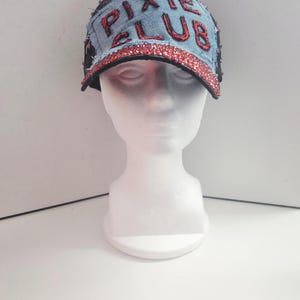 Könnte beinhalten: Eine Baseballkappe aus Jeansstoff mit den Worten "PIXIE CLUB" in Rot gestickt. Der Rand und der Text sind mit rotem Glitzer umrandet. Die Kappe wird auf einem weißen Schaufensterkopf präsentiert.