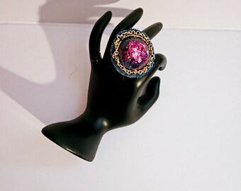 Peut inclure: Une bague imposante avec une base en jean, une bordure en chaîne dorée et un dôme transparent rempli de paillettes roses. La bague est présentée sur un présentoir noir en forme de main, mettant en valeur son design audacieux et son éclat.