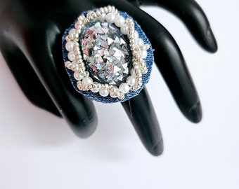 Peut inclure: Une bague en jean avec une bordure de perles argentées et blanches et une grande pierre ovale irisée au centre.