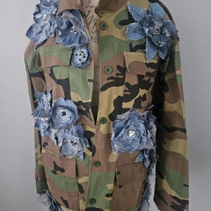 May include: Camouflage jacket adorned with denim and rhinestone floral embellishments. The jacket features a classic military-style design with button closures and pockets. The blue denim flowers are strategically placed across the front and sleeves, adding a unique, artistic touch.