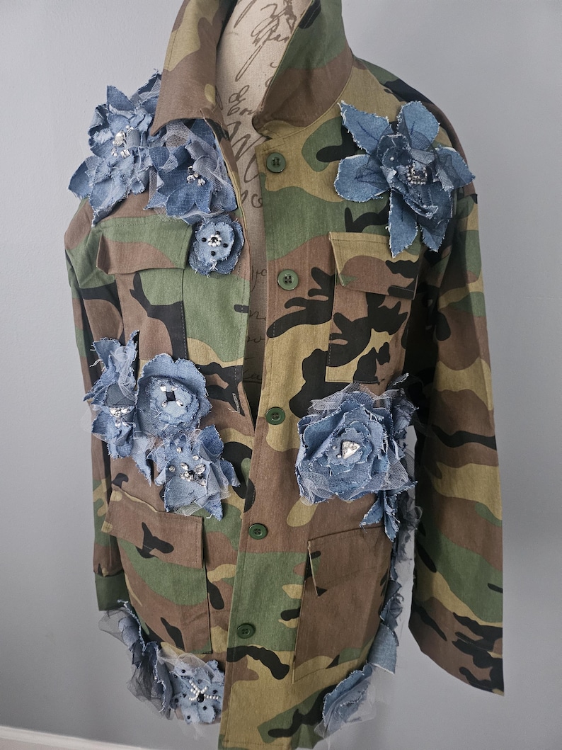 May include: Camouflage jacket adorned with denim and rhinestone floral embellishments. The jacket features a classic military-style design with button closures and pockets. The blue denim flowers are strategically placed across the front and sleeves, adding a unique, artistic touch.