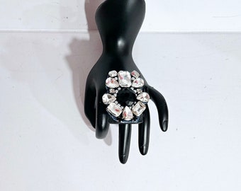 Pode incluir: Um anel preto com um grande aglomerado de strass transparentes no centro. Os strass são dispostos em um padrão circular e têm um design ligeiramente elevado e facetado. O anel é montado em uma banda de anel preta.