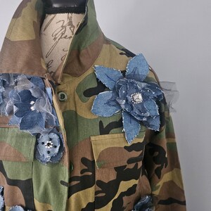 May include: Camouflage jacket adorned with denim and tulle flower embellishments. The jacket features a classic military-style design with a button-up front, pockets, and a collared neckline. The denim flowers are in various sizes and are embellished with rhinestones.