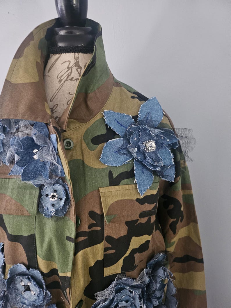 May include: Camouflage jacket adorned with denim and tulle flower embellishments. The jacket features a classic military-style design with a button-up front, pockets, and a collared neckline. The denim flowers are in various sizes and are embellished with rhinestones.