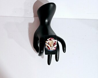 Peut inclure: Une bague avec une grande pierre précieuse facetée transparente entourée de perles rouges, blanches et bleues.