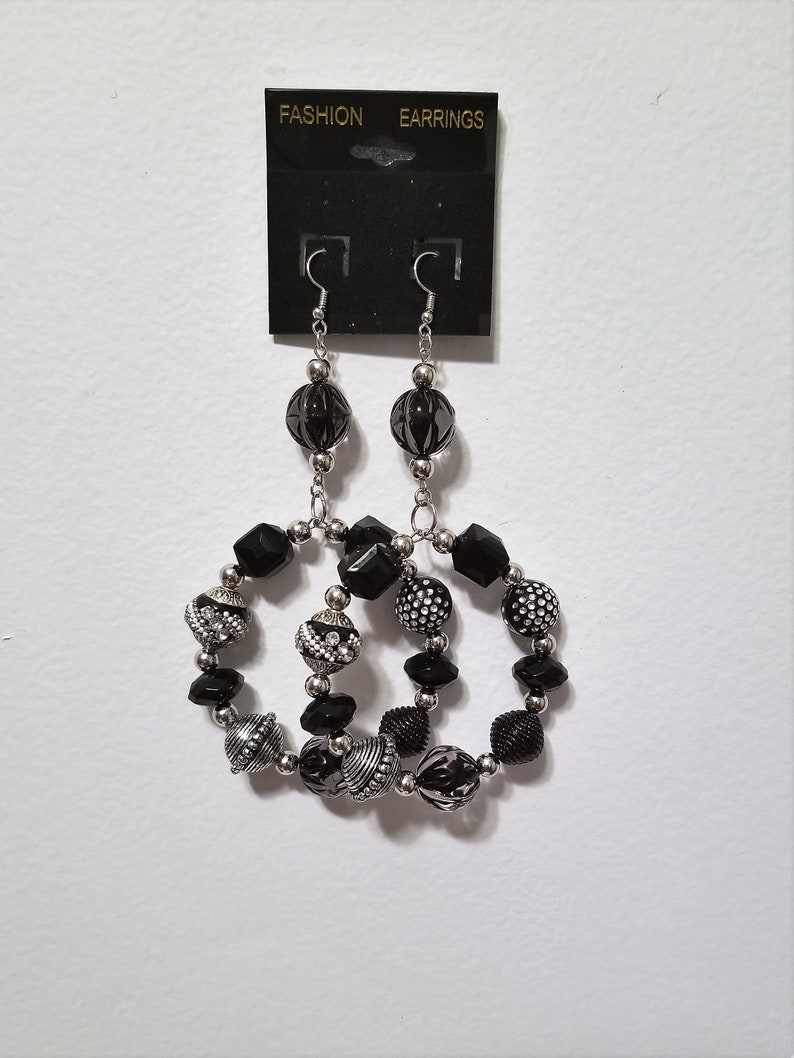 May include: Black and silver beaded hoop earrings with a silver hook closure. The earrings are on a black card with the words "FASHION" and "EARRINGS" printed on it.