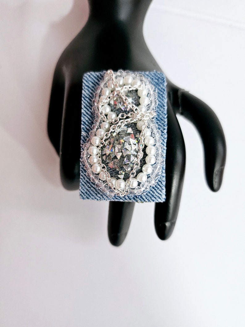 Pode incluir: Um anel de ganga com uma corrente prateada e detalhes de p&eacute;rolas. O anel apresenta uma pedra grande, brilhante e iridescente no centro.