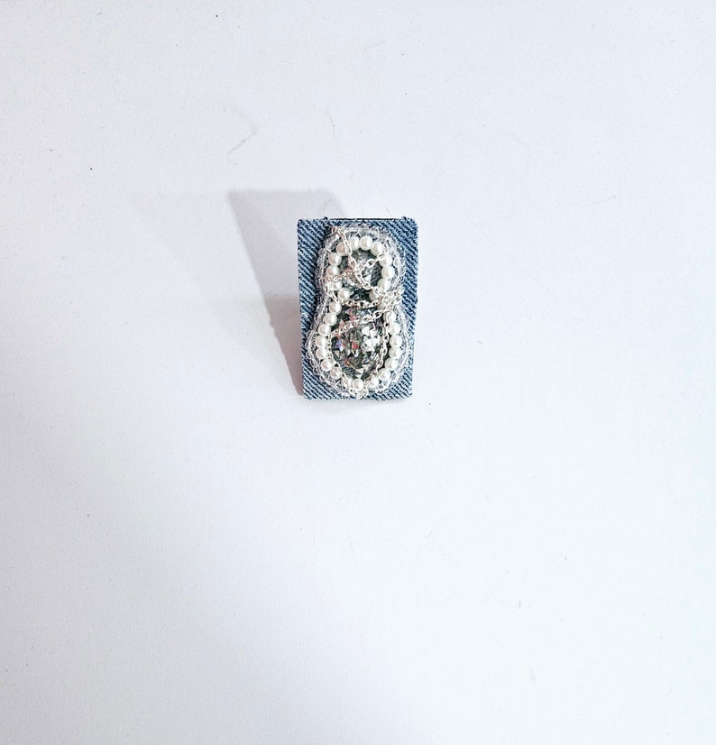 Pode incluir: Um remendo de ganga com um design de contas brancas e prateadas em forma de oito.