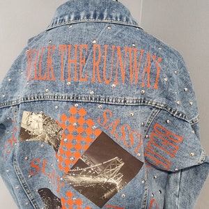 May include: A light blue denim jacket with silver studs and orange text that says "Walk the Runway", "Sassy", "Girl", and "Fashion". The jacket has a graphic print of a bridge and a checkered pattern.