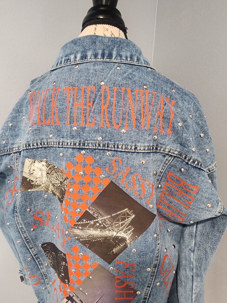 May include: A light blue denim jacket with silver studs and orange text that says "Walk the Runway", "Sassy", "Girl", and "Fashion". The jacket has a graphic print of a bridge and a checkered pattern.
