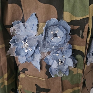 May include: Camouflage jacket with denim flower embellishments. The jacket features a classic military-style design in shades of green, brown, and black. Denim flowers with silver accents and tulle are sewn onto the jacket, adding a unique, artistic touch.