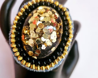 Puede incluir: Un anillo dorado y negro con un gran cabujón de resina de forma ovalada lleno de purpurina dorada.