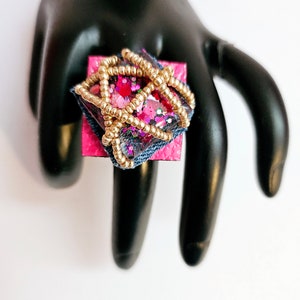 Pode incluir: Um anel quadrado com uma base rosa e uma parte superior de resina transparente. A resina est&aacute; cheia de glitter rosa, vermelho e dourado e &eacute; cercada por um fio dourado. O anel est&aacute; em um expositor de anel preto.