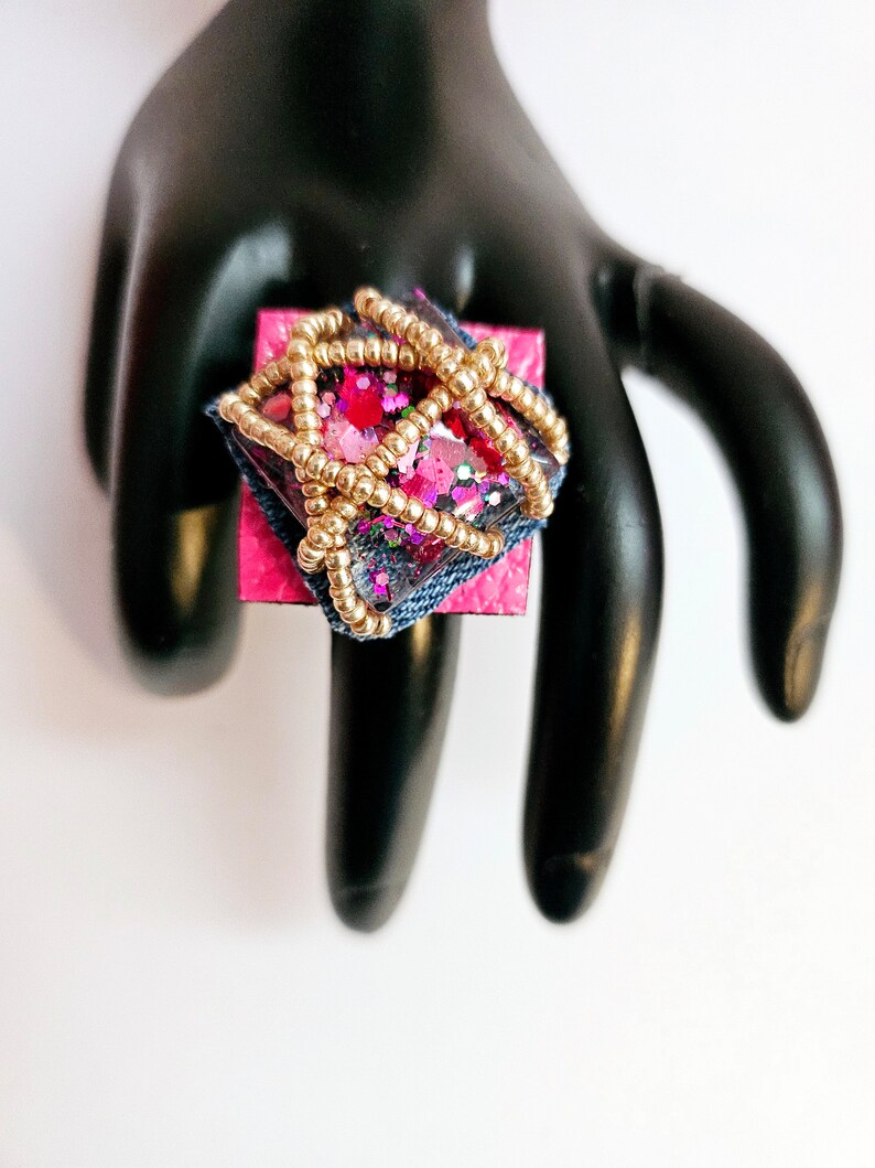 Pode incluir: Um anel quadrado com uma base rosa e uma parte superior de resina transparente. A resina est&aacute; cheia de glitter rosa, vermelho e dourado e &eacute; cercada por um fio dourado. O anel est&aacute; em um expositor de anel preto.