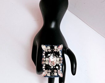 Peut inclure: Une bague rectangulaire avec une base en jean bleu et un grand cristal transparent entouré de petits cristaux noirs et transparents.
