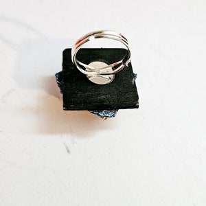 Pode incluir: Um anel ajust&aacute;vel de prata com um design de tecido azul e branco. O anel &eacute; exibido em uma base quadrada preta.