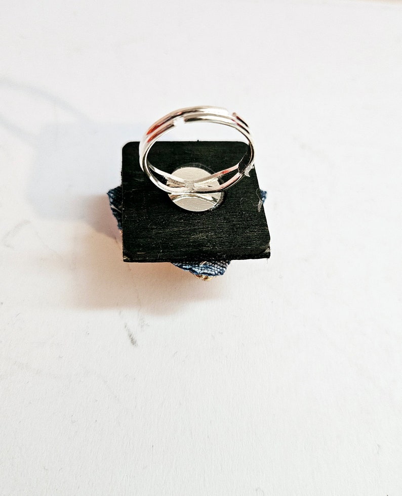 Pode incluir: Um anel ajust&aacute;vel de prata com um design de tecido azul e branco. O anel &eacute; exibido em uma base quadrada preta.