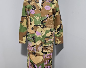 Op de afbeelding: Een lange, groene camouflagejas met een ritssluiting en zakken. De jas heeft verschillende ontwerpen van roze en gouden glitter, waaronder harten, sterren, lippen en de woorden "New York", "Walk the Runway", "High Fashion", "I Love NY" en "Fashion is Everything".