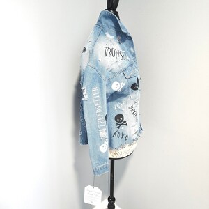 Puede incluir: Una chaqueta vaquera azul claro con detalles desgastados y varios dise&ntilde;os gr&aacute;ficos en blanco y negro, incluyendo calaveras, corazones y las palabras "Promise", "Trendsetter" y "XOXO".