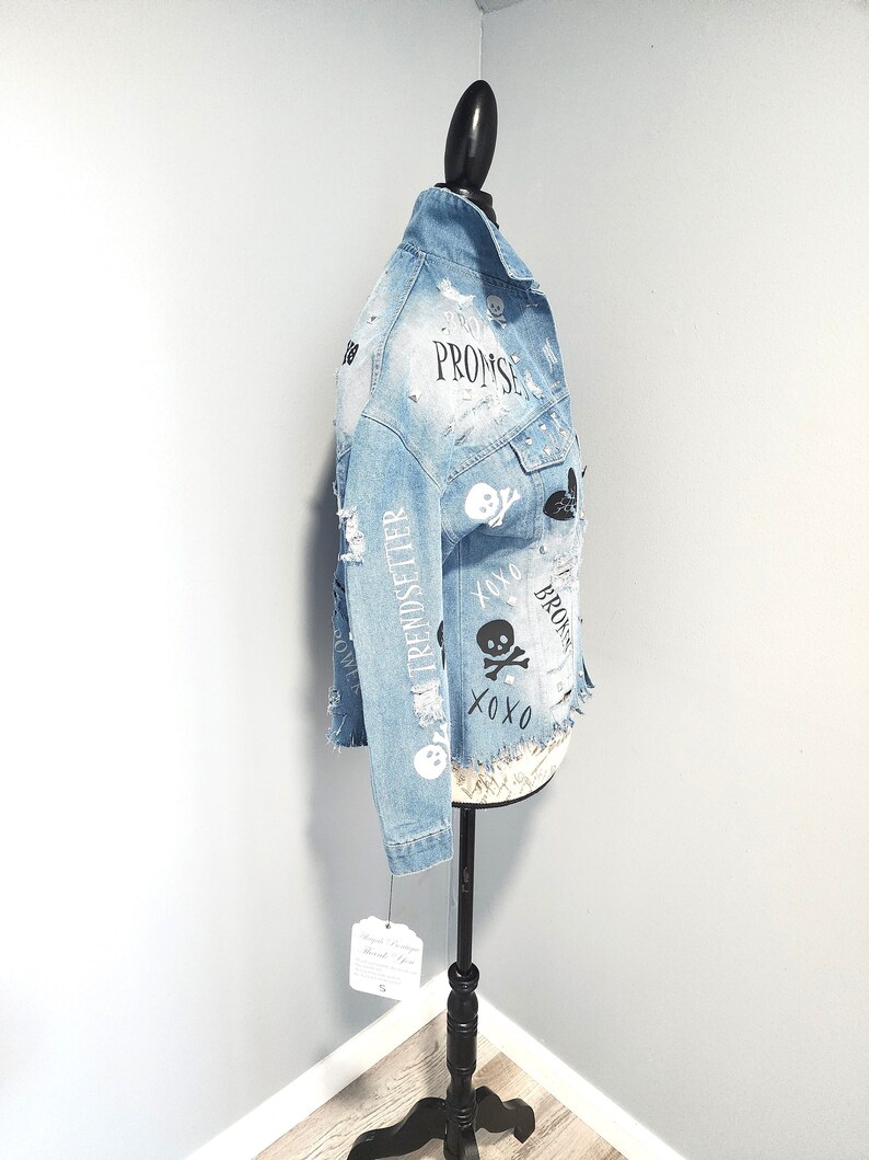 Puede incluir: Una chaqueta vaquera azul claro con detalles desgastados y varios dise&ntilde;os gr&aacute;ficos en blanco y negro, incluyendo calaveras, corazones y las palabras "Promise", "Trendsetter" y "XOXO".