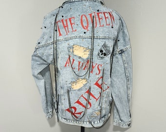 Puede incluir: Chaqueta vaquera azul claro con texto rojo que dice "The Queen Always Rule" y una calavera con un gráfico de corona. La chaqueta tiene tachuelas y cadenas plateadas.