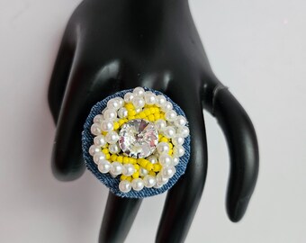 Peut inclure: Une grande bague statement en tissu denim, perles blanches et perles jaunes. La bague présente un grand strass transparent au centre.