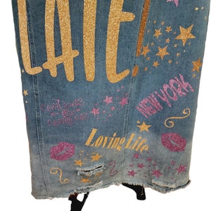 May include: A blue denim jacket with gold glitter lettering that says "LATE!" and "Loving Life" and pink glitter lettering that says "NEW YORK" and has pink glitter stars and lips.