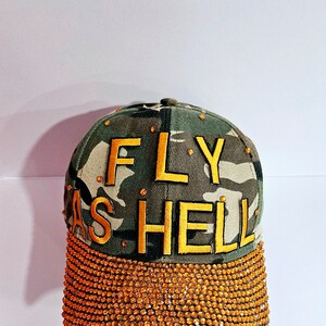 Op de afbeelding: Een camouflage baseballpet met een gouden geborduurde tekst "FLY AS HELL". De rand is bedekt met gouden strass.