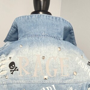 Puede incluir: Una chaqueta vaquera azul claro con tachuelas plateadas y detalles desgastados. La parte posterior de la chaqueta presenta la palabra "RAGE" en letras blancas, una calavera y huesos cruzados, y el texto "FASHION" en letras blancas.