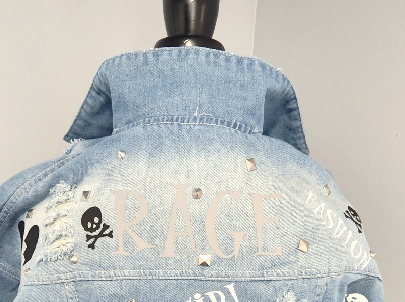 Puede incluir: Una chaqueta vaquera azul claro con tachuelas plateadas y detalles desgastados. La parte posterior de la chaqueta presenta la palabra "RAGE" en letras blancas, una calavera y huesos cruzados, y el texto "FASHION" en letras blancas.
