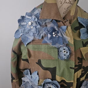 May include: Camouflage military jacket adorned with denim and tulle flowers. The jacket features a classic camouflage pattern in shades of brown, green, and black. Denim flowers with rhinestone centers are attached to the jacket, adding a touch of femininity to the rugged style.