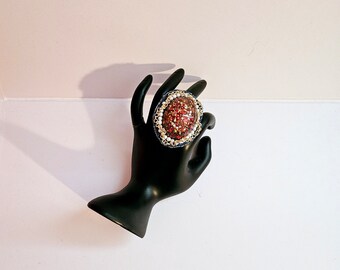 May include: A large, oval-shaped ring with a red and gold glitter resin inlay. The ring is surrounded by a silver-toned metal frame with white pearls and black beads.