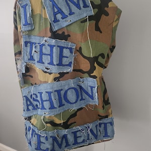 May include: Camouflage vest with denim patches spelling "I AM THE FASHION MOVEMENT" in blue thread. The vest has a classic military-inspired pattern in shades of green, brown, and black.