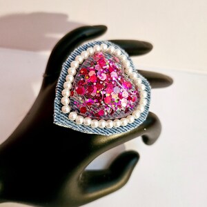 K&ouml;nnte beinhalten: Ein herzf&ouml;rmiger Ring aus Harz mit eingebetteten pinkfarbenen und roten Glitzer. Der Ring ist von einem wei&szlig;en Perlenbesatz und einer Basis aus Jeansstoff umgeben.