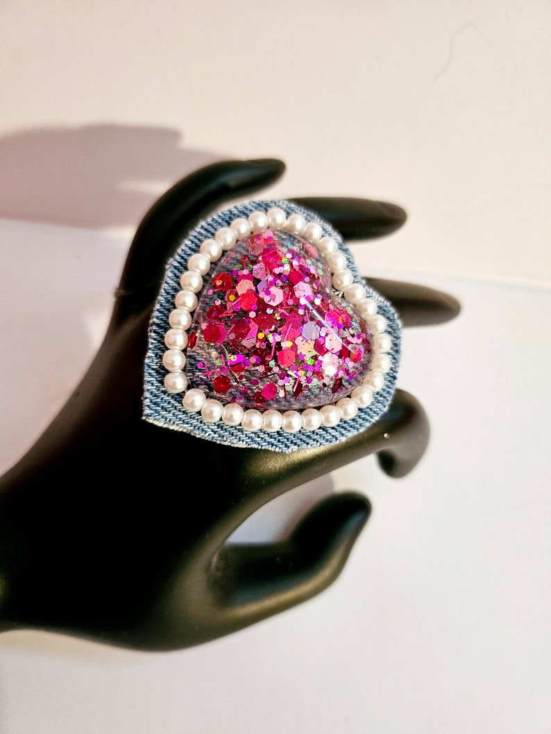 K&ouml;nnte beinhalten: Ein herzf&ouml;rmiger Ring aus Harz mit eingebetteten pinkfarbenen und roten Glitzer. Der Ring ist von einem wei&szlig;en Perlenbesatz und einer Basis aus Jeansstoff umgeben.