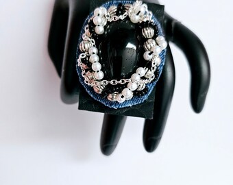 Peut inclure: Une bague noire avec une grosse pierre noire entourée de chaînes argentées, de perles blanches et de perles noires. La bague est posée sur une base en jean bleu.