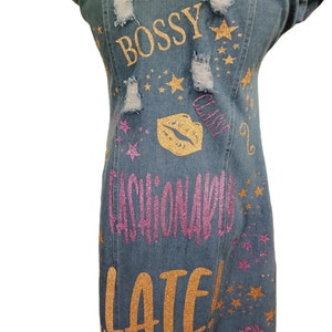 May include: A blue denim dress with gold and pink glitter lettering that says "Bossy, Class, Fashionably Late". The dress has several distressed areas and is decorated with gold glitter stars.