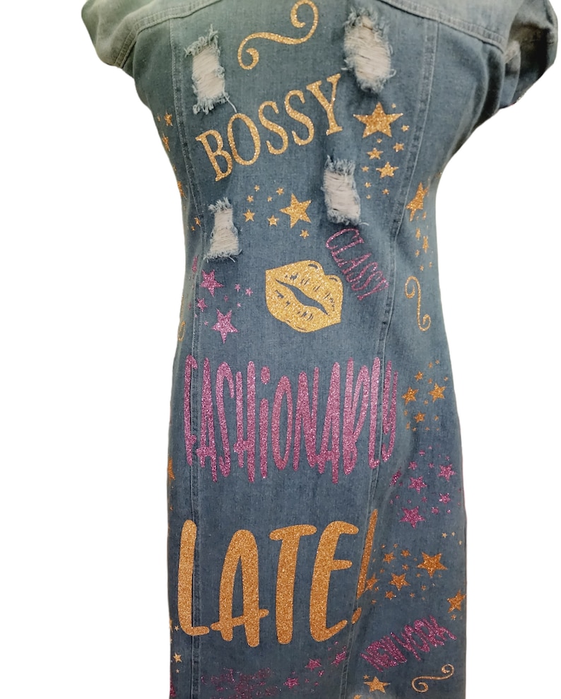 May include: A blue denim dress with gold and pink glitter lettering that says "Bossy, Class, Fashionably Late". The dress has several distressed areas and is decorated with gold glitter stars.