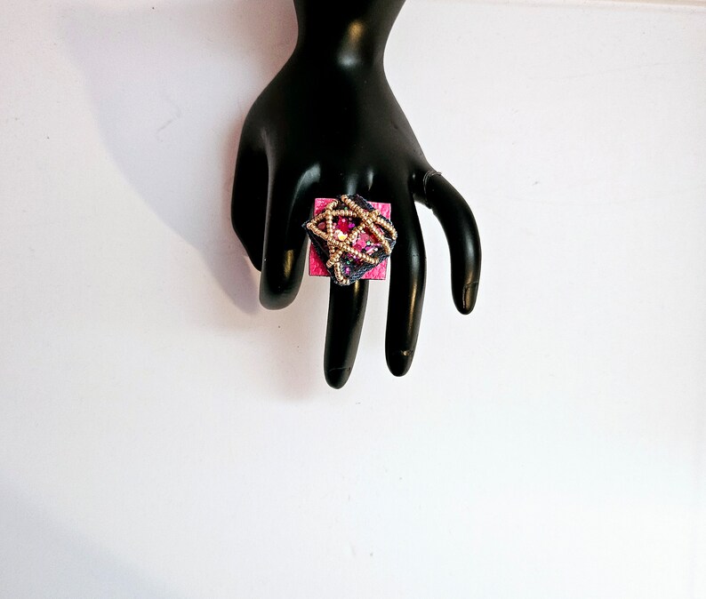 Pode incluir: Um anel quadrado com fundo rosa e design de contas douradas e azuis.