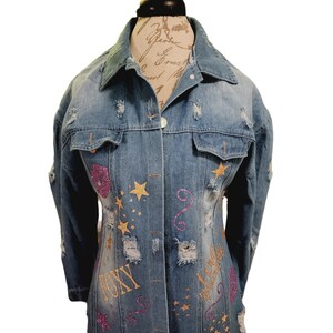 May include: A long denim jacket with distressed details and gold glitter stars and writing. The writing includes the words "Diva", "Queen", and "Tony".