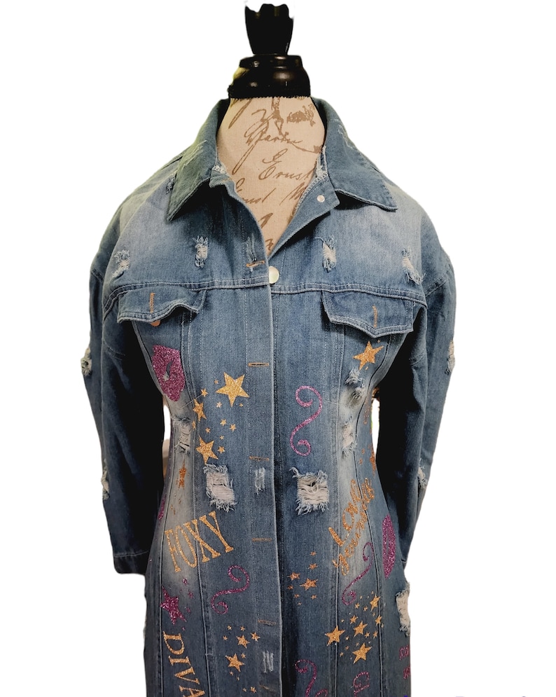 May include: A long denim jacket with distressed details and gold glitter stars and writing. The writing includes the words "Diva", "Queen", and "Tony".