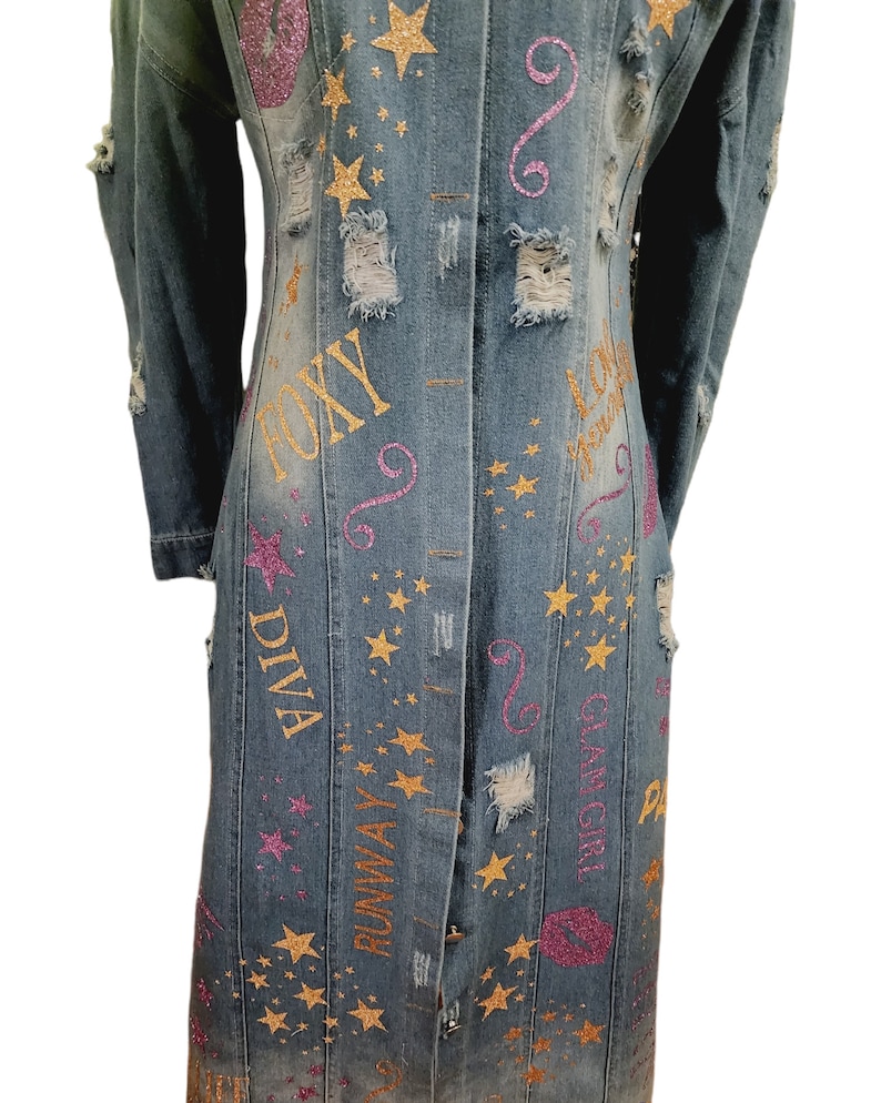 May include: A long denim jacket with distressed details and gold and pink glitter lettering. The lettering includes the words "Foxy Diva", "Runway", "Glam Girl", and "Love You".