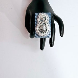 Pode incluir: Um anel quadrado azul denim com um design de contas prateadas e brancas em forma de n&uacute;mero oito.