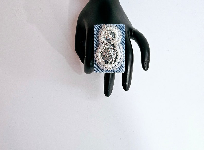 Pode incluir: Um anel quadrado azul denim com um design de contas prateadas e brancas em forma de n&uacute;mero oito.