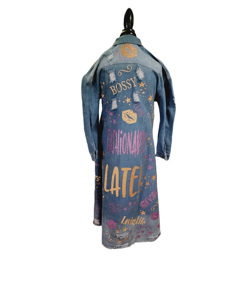 May include: A long denim jacket with distressed details and glitter accents. The jacket features the words "Bossy", "Fashionably Late", "New York", and "Loving Life" in pink and gold glitter.
