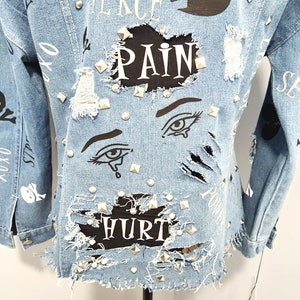 Puede incluir: Una chaqueta vaquera azul claro con detalles desgastados, tachuelas y parches negros con las palabras "Fierce", "Pain" y "Hurt" impresas en ellos.