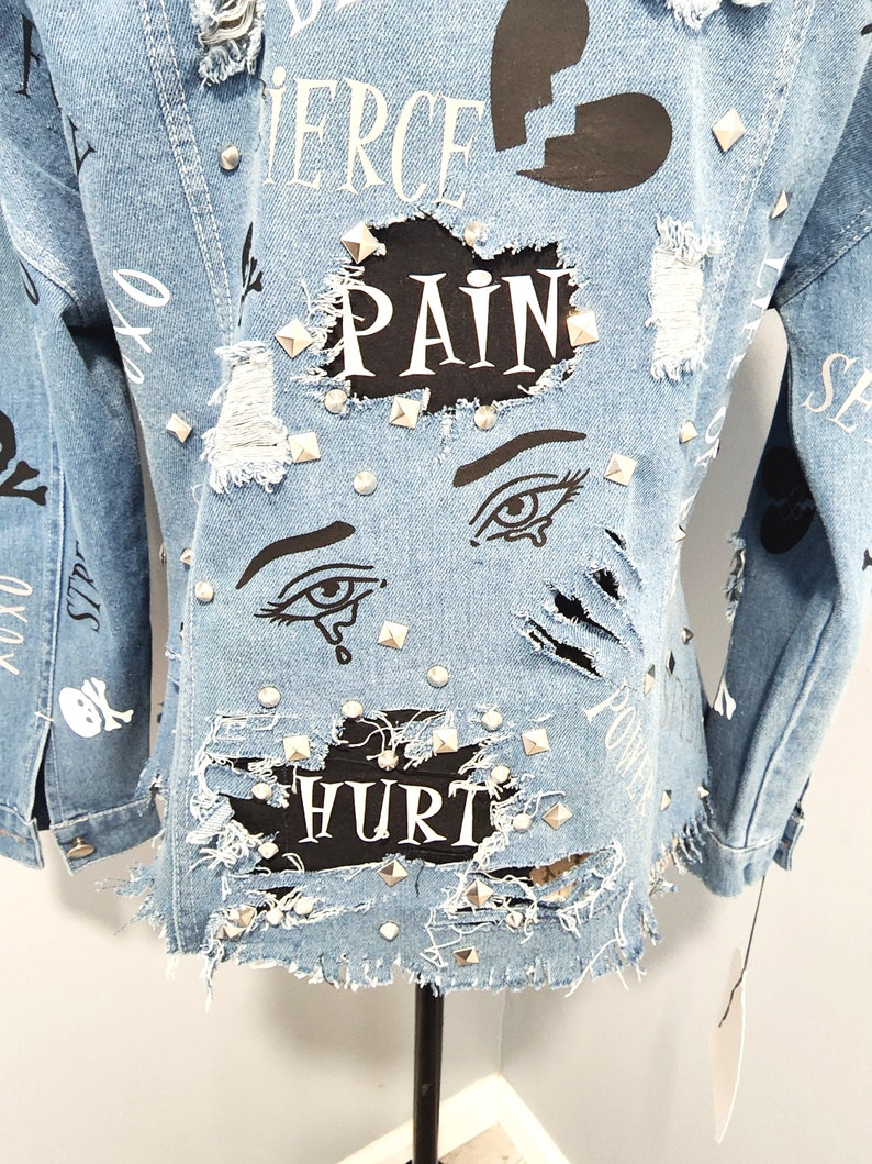 Puede incluir: Una chaqueta vaquera azul claro con detalles desgastados, tachuelas y parches negros con las palabras "Fierce", "Pain" y "Hurt" impresas en ellos.