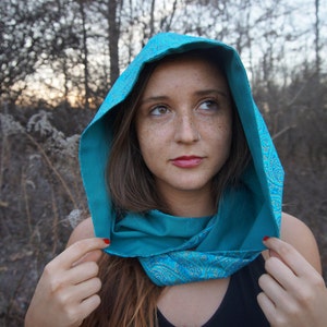 May include: A woman wearing a turquoise hooded scarf with a paisley pattern. The scarf is tied around her neck and the hood is pulled up over her head. She is looking off to the side.