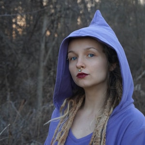 Puede incluir: Una mujer con largas rastas rubias lleva una sudadera con capucha morada. Tiene un piercing en la nariz y maquillaje de ojos oscuro.
