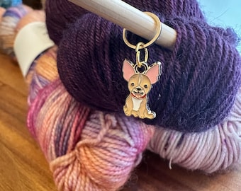 May include: A gold-colored metal yarn marker featuring a cartoon chihuahua with pink ears and a white collar. The marker is attached to a wooden knitting needle and is surrounded by two skeins of yarn, one purple and one pink and orange.